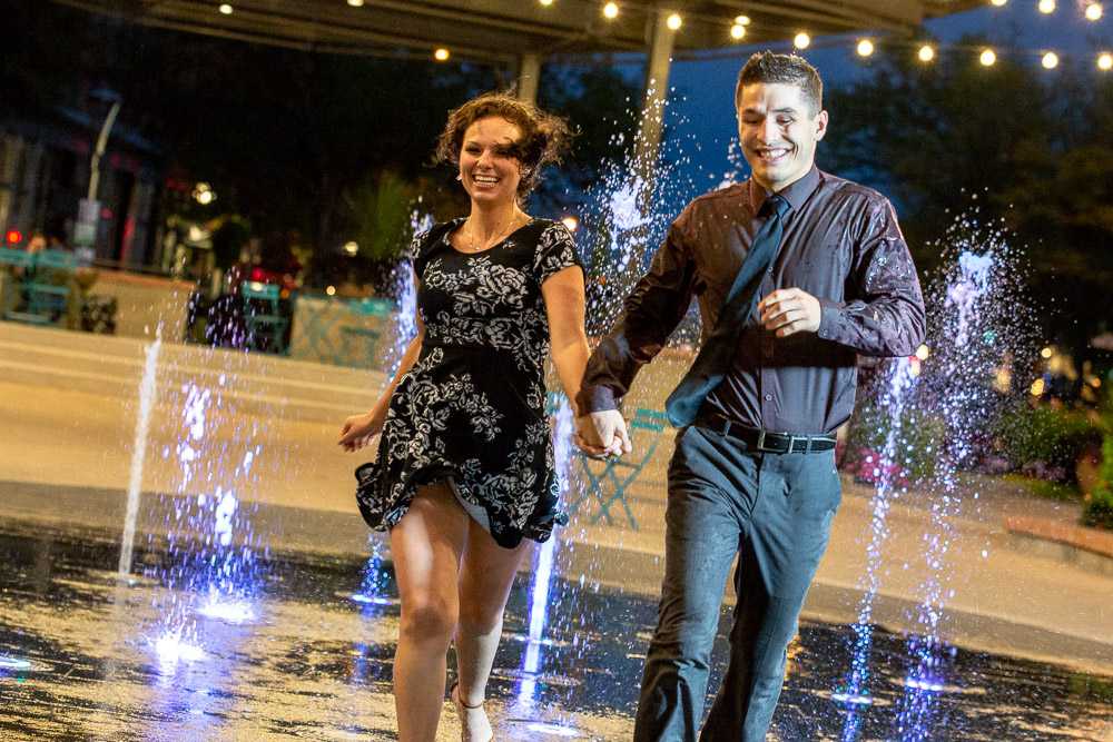 An engaged couple holds hands and runs through a fountain at night in Old Town Fort Collins.