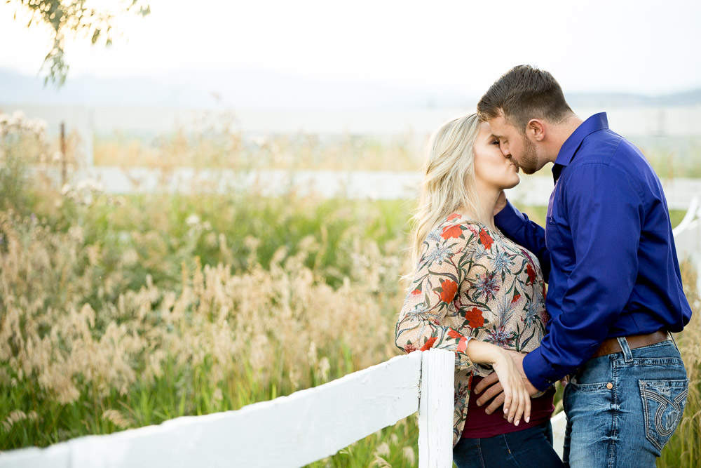 A couple leaning against a white fence kissing in a field at their outdoor Fort Collins engagement photo session.